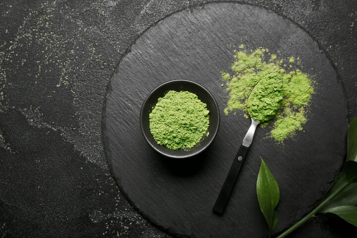 Matcha powder in black bowl with spoon and leaves on dark slate