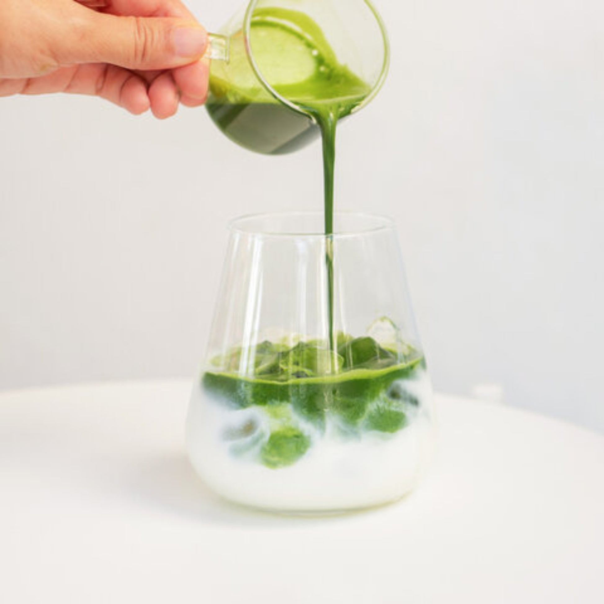 Pouring BENBU Matcha into iced milk to make an iced matcha latte in a clear glass on white background