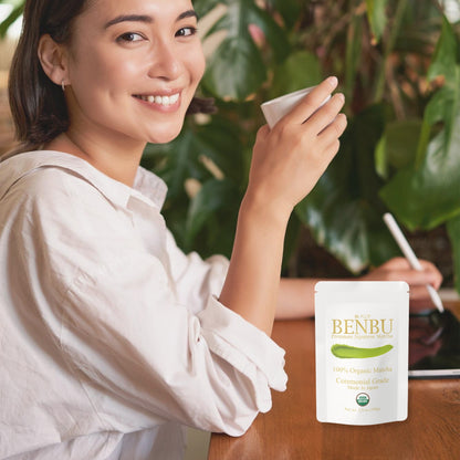 Woman drinking BENBU Organic Matcha with 100 g bag on wooden table
